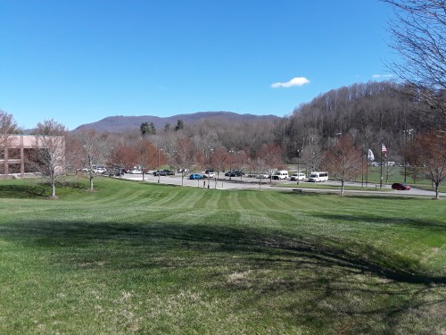 A grassy area with trees and a clear blue sky, overlooking a road with parked cars and distant mountains.