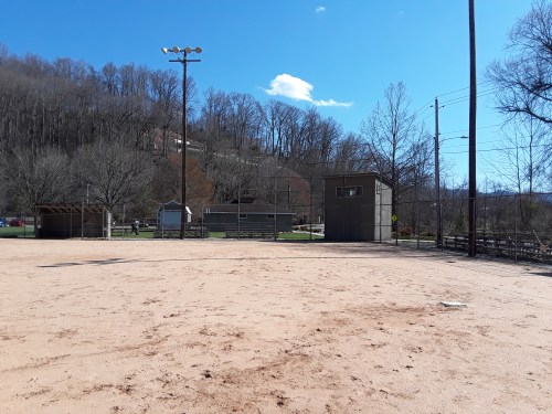 A sandy baseball field with a backdrop of trees and a clear blue sky, featuring a dugout and a scoreboard.