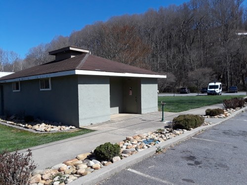 A small, gray building with a brown roof, surrounded by grass and rocks, set against a wooded hillside.