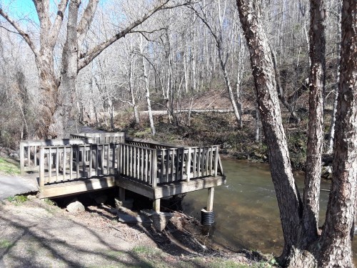 A wooden deck by a calm stream, surrounded by bare trees and a path in a serene natural setting.
