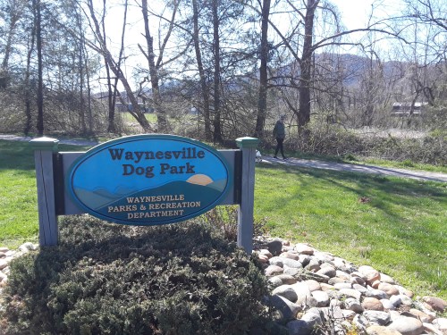 Sign for Waynesville Dog Park surrounded by greenery, with a person walking in the background on a sunny day.