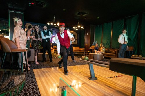 A man in a red hat bowls while a group of elegantly dressed friends watches in a stylish lounge setting.