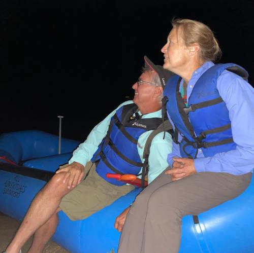 Two people in life jackets sit on a raft at night, looking up and smiling, with a dark background.