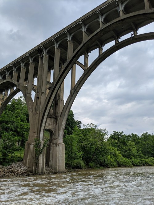 A large concrete bridge arches over a river, surrounded by lush green trees under a cloudy sky.