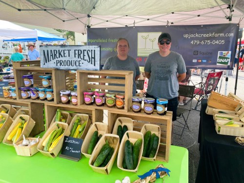 Two vendors stand behind a table displaying fresh produce and jars of goods at a farmers market.