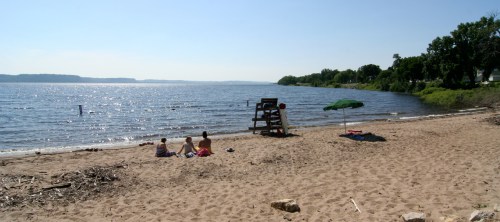 Two people sit on a sandy beach by a calm lake, with a lifeguard chair and an umbrella nearby under a clear blue sky.
