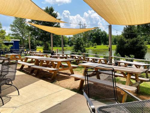 Outdoor seating area with picnic tables and shade sails by a pond, surrounded by trees and blue skies.