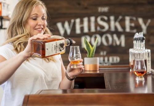 A woman pours whiskey into a glass at a bar with a "Whiskey Room" sign in the background.
