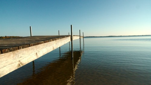 A wooden dock extends into calm water under a clear blue sky, reflecting the serene landscape.