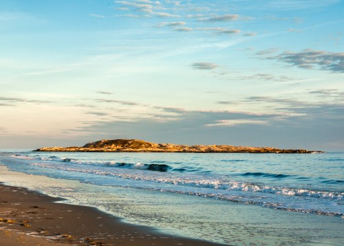 A serene beach scene with gentle waves lapping at the shore and a small island in the distance under a colorful sky.