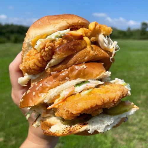 A hand holding a stacked fried chicken sandwich with lettuce, pickles, and sauces against a green field backdrop.