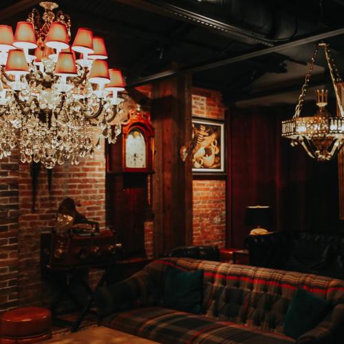 Cozy interior featuring a vintage chandelier, brick walls, a classic clock, and a plush sofa with plaid upholstery.