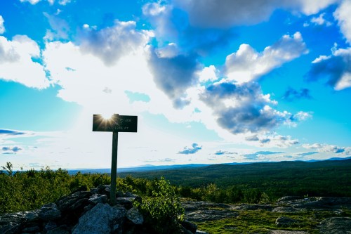 A scenic view from a mountain top with a signpost, bright sun rays, and a blue sky dotted with clouds.