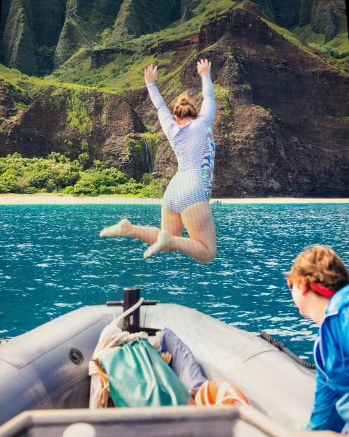 A person jumps off a boat into the blue water, with lush green cliffs and a beach in the background.