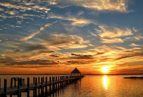 A serene sunset over a calm lake, with a wooden pier leading to a small gazebo and colorful clouds in the sky.