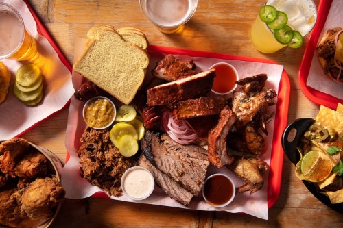 A vibrant platter of assorted barbecue meats, pickles, bread, and sauces, served with drinks on a wooden table.