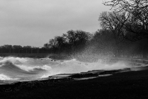 Waves crash against a rocky shoreline on a cloudy day, with trees in the background and mist rising from the water.
