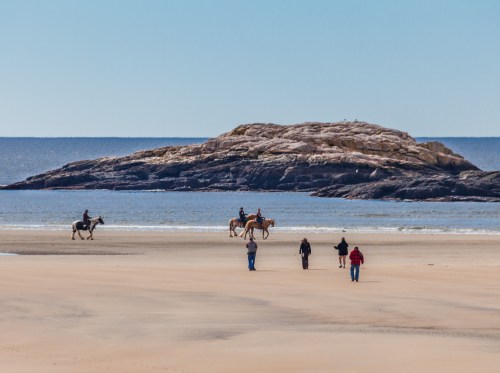 Two horseback riders on a beach, with people walking along the shore and a rocky island in the background.