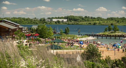 A sunny beach scene with people enjoying the water, surrounded by greenery and a calm lake under a blue sky.