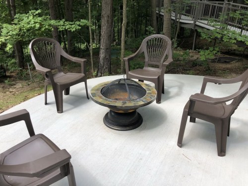 A circular patio with four brown chairs surrounding a stone fire pit, set in a wooded area.