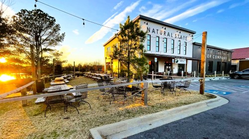A cozy restaurant with outdoor seating, surrounded by trees, at sunset with a clear sky.