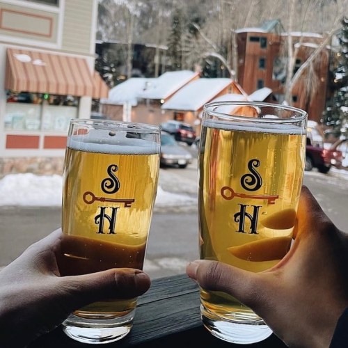 Two hands hold glasses of beer with a logo, set against a snowy outdoor scene with buildings in the background.