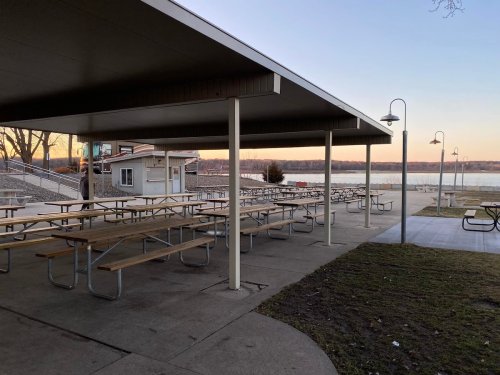 A covered picnic area with empty wooden tables, overlooking a river at sunset.