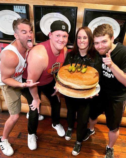 Four people pose with a giant burger topped with pickles, smiling and making playful gestures in a restaurant setting.