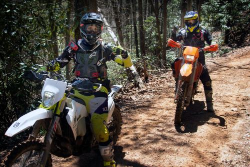 Two motocross riders on dirt bikes in a forested area, one giving a thumbs-up while the other stands beside their bike.