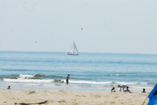 A sailboat glides across a calm sea, with beachgoers enjoying the sandy shore in the foreground.