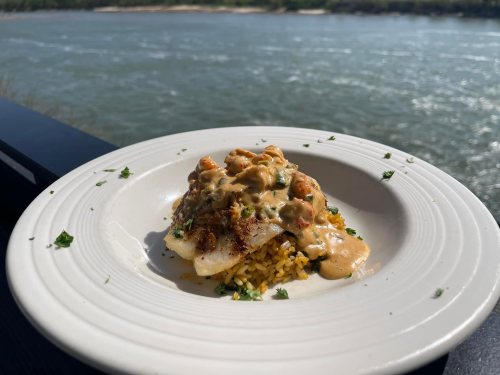 A plate of seasoned fish topped with creamy sauce, served over rice, with a scenic river view in the background.