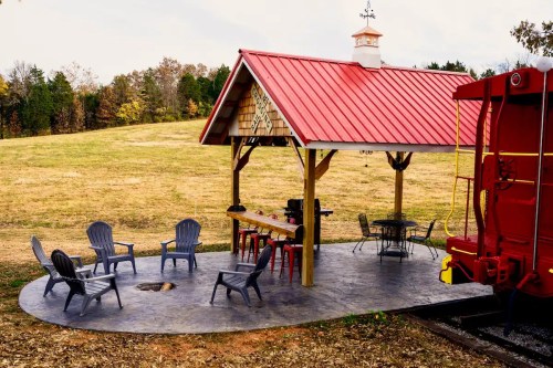 A cozy outdoor patio with a red-roofed shelter, seating, and a vintage train car nearby, set in a grassy landscape.