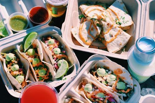 A vibrant spread of tacos, quesadillas, and drinks in takeout containers, garnished with lime and fresh toppings.