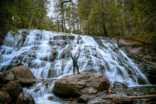 A person stands on a rock in front of a cascading waterfall, surrounded by lush green trees.