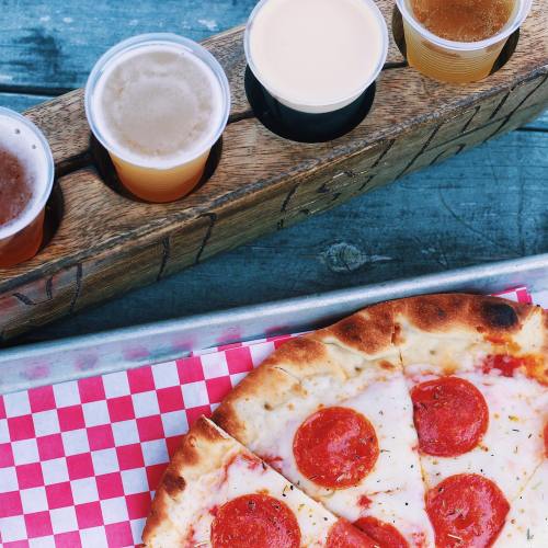 A wooden flight of beer samples next to a pepperoni pizza on a checkered paper.