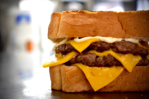 A close-up of a hearty sandwich with two beef patties, melted cheese, and mayonnaise between thick slices of bread.