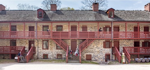 Historic stone building with red balconies, featuring people on the stairs and in front, surrounded by trees.
