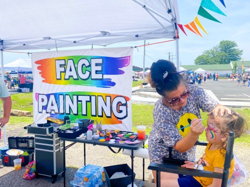 A face painter applies colorful designs to a child's face at an outdoor event, with a "Face Painting" sign in the background.