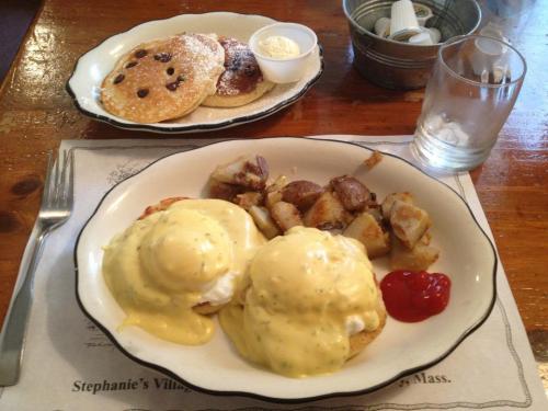 A plate of eggs Benedict with potatoes and ketchup, alongside a bowl of pancakes with chocolate chips and a small cup of cream.