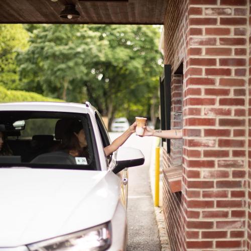 A person in a car receives a drink from a server at a drive-thru window surrounded by greenery.