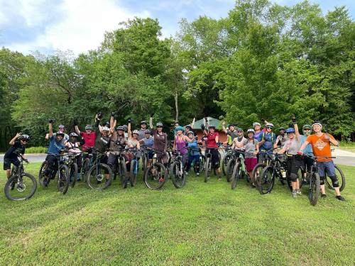 A large group of people on mountain bikes, smiling and celebrating outdoors in a green park setting.