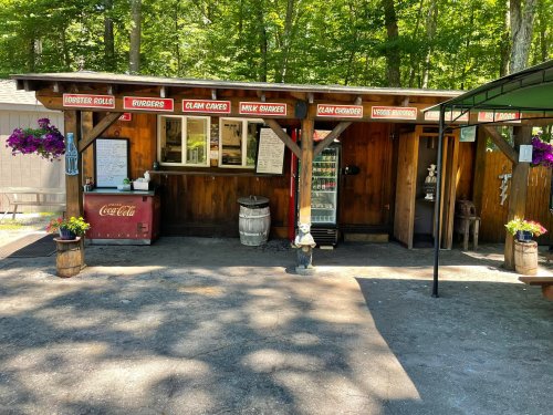 A rustic food stand with a wooden structure, offering lobster rolls, burgers, and clam cakes, surrounded by greenery.