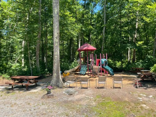 A playground with slides and climbing structures surrounded by trees, with picnic tables and chairs nearby.