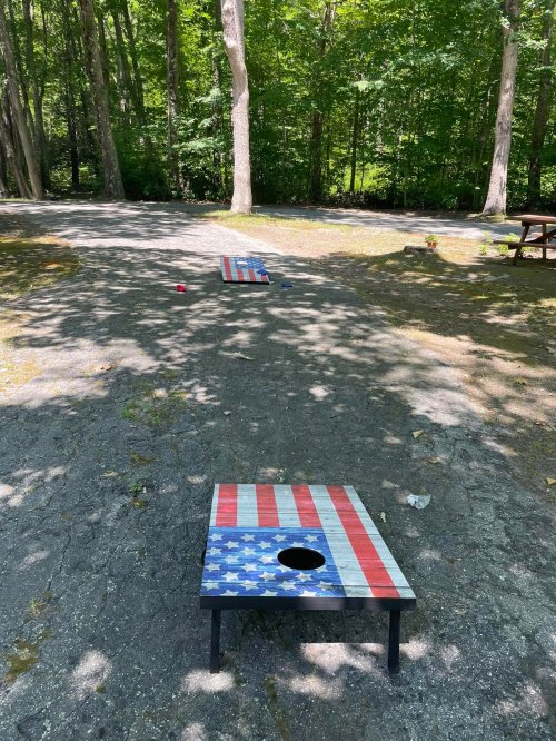 Two cornhole boards with American flag designs set on a gravel path in a wooded area, surrounded by trees.