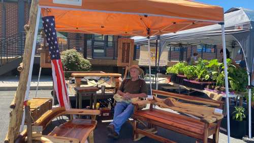 A man sits under an orange tent at a market, surrounded by wooden furniture and plants, with an American flag nearby.