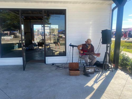 A musician plays guitar and sings outside a building, with a crowd visible in the background.