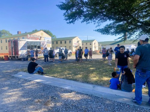 A food truck is parked near a group of people gathered outside, with buildings in the background and trees providing shade.