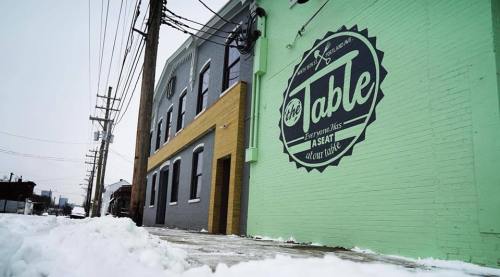 A colorful mural on a building reads "Everyone has a seat at our table," with snow on the ground and power lines nearby.