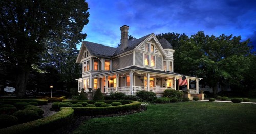 A charming Victorian house with a porch, surrounded by manicured gardens, illuminated at dusk.