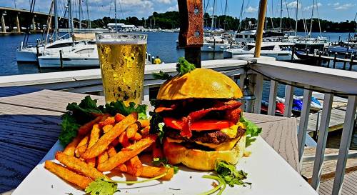 A delicious burger with bacon and tomato, served with sweet potato fries and a cold beer, overlooking a marina.